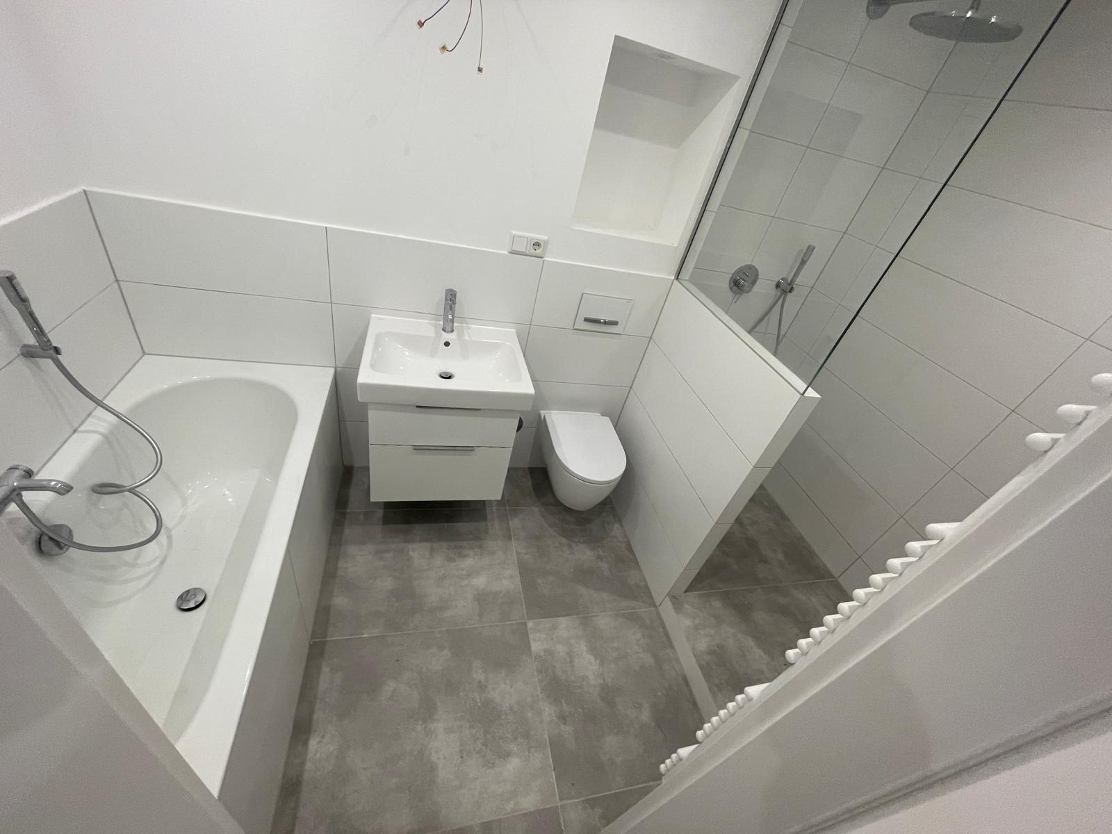 Modern saniertes Badezimmer mit Badewanne, Dusche mit Glaswand, wandhängendem WC und weißem Waschtisch auf großen grauen Bodenfliesen