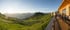 Panoramablick auf eine Berghütte mit Terrasse und Tischen im Vordergrund, dahinter grüne Hügel und Berge.