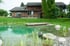 Ein Schwimmteich mit Holzsteg und Wasserpflanzen vor einem rustikalen Holzhaus mit bewachsener Fassade.