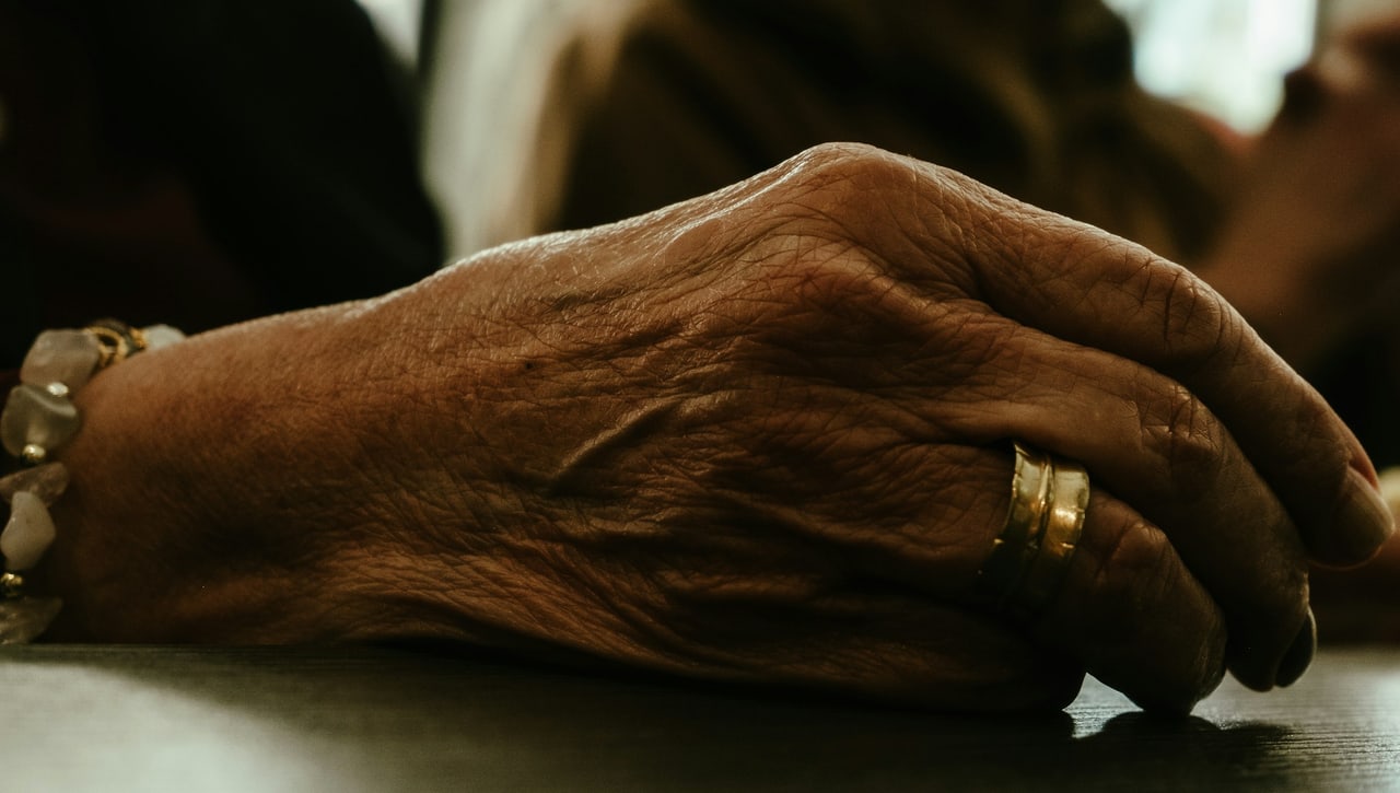 Die faltige Hand eines älteren Menschen mit Ring und Armband ruht in warmem, gedämpftem Licht auf einer Tischkante.