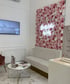 Warteraum mit rosa Blumenwand, Leuchtschild "Beauty Atelier by Filiz", weißer Couch, Marmortisch, TV und Broschüren.