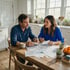 A man and woman sit at a wooden kitchen table reviewing financial papers and a calendar over coffee.