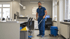 Ein Mann in blauer Uniform wischt einen Büroboden mit einem Mopp, neben ihm ein blauer Eimer.