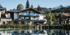 Modernes Holzhaus mit Glasbalkonen spiegelt sich in einem ruhigen See, umgeben von grünen Bäumen und Bergen.