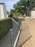 Ein grauer Doppelstabmattenzaun verläuft auf einem Betonsockel zwischen einem gepflasterten Weg und einer sandigen Fläche.