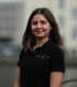 Lächelnde Frau mit braunen Haaren trägt ein schwarzes Poloshirt mit dem Logo Schmelter & Schütt vor hellem Hintergrund.