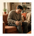 An older man sits on a brown sofa wincing while clutching his knee in a living room.