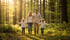 Eine Familie mit zwei kleinen Kindern geht Hand in Hand auf einem Waldweg, umgeben von hohen Bäumen und Farnen.
