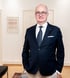 Ein lächelnder Mann in Anzug und Brille steht in einem hellen Flur mit Sofa und Bildern.