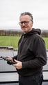 Ein lächelnder Mann mit Brille und schwarzer Kapuzenjacke hält ein Tablet auf einem Dach mit Solarmodulen.