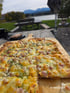 Eine Käse-Schinken-Pizza auf einem Holzbrett mit Blick auf einen See und Berge, Text "HAP 19. Oktober 2025".