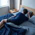 A man in a blue t-shirt sleeps on a bed with navy linens next to a nightstand with a digital clock reading 5:37.