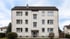 Dreistöckiges Mehrfamilienhaus mit weißer Fassade, Satteldach und zentralem Eingangsbereich unter bewölktem Himmel.