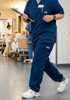 Medical professional in blue scrubs and white clogs walks down a hospital hallway holding a clipboard.