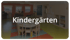 Ein heller Gruppenraum im Kindergarten mit Holzstühlen, Tischen und Regalen sowie dem zentralen Schriftzug „Kindergärten“.