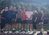 Eine Gruppe von acht Personen in Sportkleidung posiert lächelnd vor einem bewaldeten Berg im Sonnenlicht.