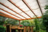 Eine hölzerne Terrassenüberdachung mit weißer Stoffbespannung über einem Holztisch mit Stühlen im Garten.