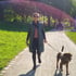 Frau in Steppmantel und Sonnenbrille geht mit braunem Hund auf einem Weg im Park.