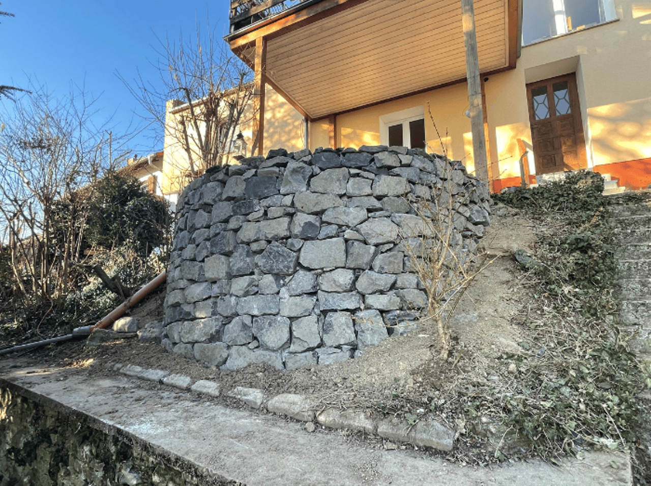 Runde Stützmauer aus Naturstein an einem Hang neben einem Haus