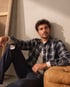 Ein sitzender Mann trägt ein dunkelblau kariertes Flanellhemd über einem weißen T-Shirt und eine dunkle Jeans.