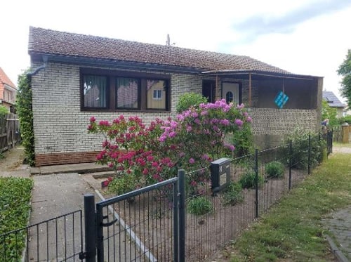 Ein kleines Einfamilienhaus mit Ziegeldach, weiß gestrichenen Ziegelwänden und einem blühenden rosa Rhododendronbusch davor.