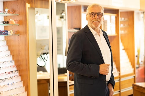 Mann im dunklen Anzug steht in einem Optikergeschäft vor Regalen mit Brillen