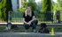 Eine Frau hockt zwischen zwei Hunden auf einer Straße; im Hintergrund ein grüner Zaun und Bäume.