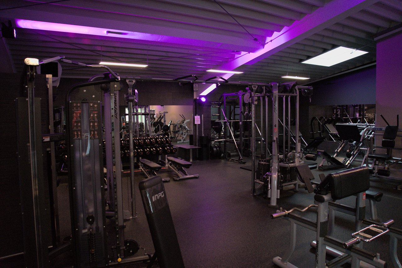 Fitnessstudio mit lila Beleuchtung und einer Vielzahl von Trainingsgeräten.