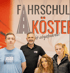 Drei Personen stehen vor einem roten Banner mit dem Logo "FAHRSCHULE KÖSTER...echt erfahren!".