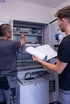 Zwei Elektriker überprüfen gemeinsam einen geöffneten Schaltschrank, einer zeigt auf Bauteile, der andere hält einen Bauplan.
