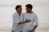 Two young men in white shirts, one with hand on the other's chest, smiling at each other by the water.