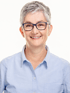 Eine lächelnde Frau mittleren Alters mit kurzen, grauen Haaren und blauer Brille, die ein hellblaues Hemd trägt.