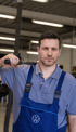 Ein Mechaniker in blauer Latzhose mit Volkswagen-Logo hält ein Werkzeug über seiner Schulter.