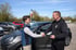 Zwei Männer schütteln sich die Hand vor einem grauen SUV; einer übergibt Autoschlüssel.