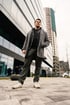 A man in a gray coat and pants stands on a paved surface outside a modern glass building.