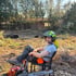 Waldarbeiter mit Schutzhelm und Husqvarna Motorsäge sitzt auf einem Stuhl im Freien vor einem bewaldeten Grundstück.