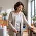 A senior woman in a cardigan uses a walking cane and a wooden shelf for support while walking through a living room.