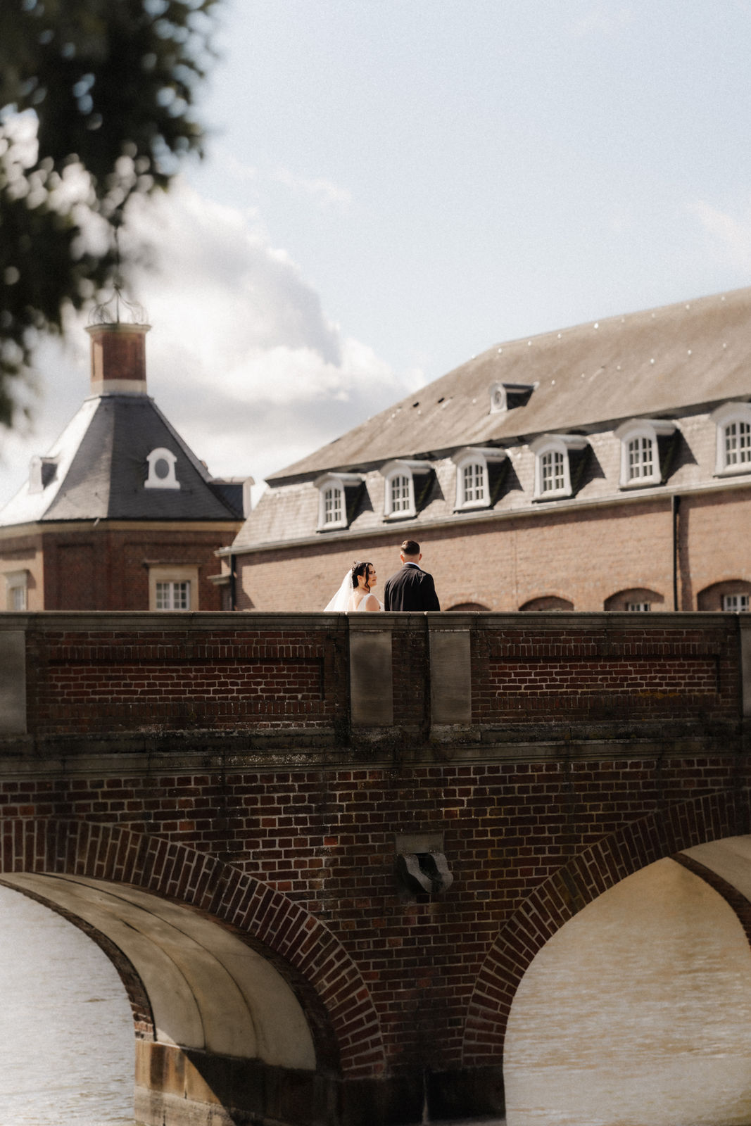 Brautpaar steht Hand in Hand auf einer Backsteinbrücke vor Schloss Nordkirchen, das sich im Wasser spiegelt.