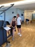 Zwei lächelnde Frauen in einem modernen Fitnessstudio neben einem Rücken-Trainingsgerät auf hellem Holzboden.