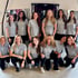 Zehn Frauen in grauen Poloshirts und schwarzen Hosen posieren lächelnd in einem Fitnessstudio.