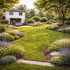 Gepflegter Garten mit üppiger, grüner Rasenfläche, umgeben von blühenden Stauden und Sträuchern, und einem weißen Haus im Hintergrund.