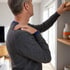A man in a grey sweater holds his shoulder in pain while reaching for a high shelf.