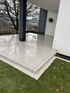Nasse, hellgraue Terrassenfliesen auf einer modernen Veranda mit dunklem Stützpfeiler und angrenzender Rasenfläche.