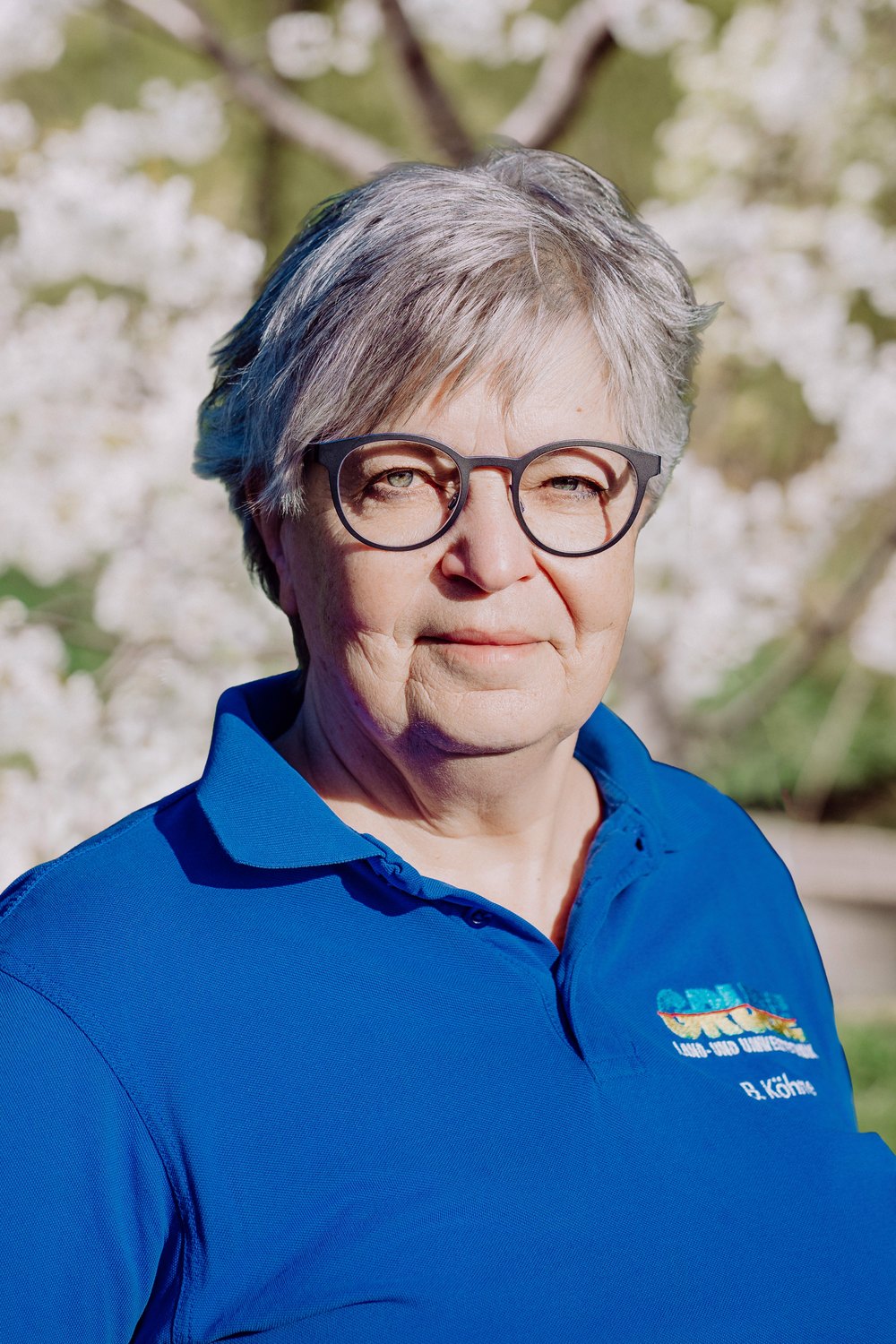 Frau mit grauen Haaren und Brille in blauem Poloshirt mit "GRÜN" Logo vor weiß blühendem Baum.
