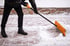 Person in schwarzer Kleidung schiebt Schnee mit einer orangefarbenen Schaufel von einem gepflasterten Weg.