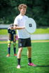Ein junger Mann im Fußballtrikot hält ein weißes rundes Schild mit der Aufschrift „TSA TRAINING AID“.
