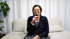 Frau mit Brille, dunkelblauem Blazer und gemustertem Oberteil sitzt auf Sofa und hält Mikrofon.