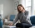 A woman in a gray sweater sits on a couch, wincing while holding her hand to her neck in a living room.