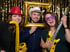 Drei lachende Erwachsene mit Party-Requisiten vor glitzerndem Hintergrund: roter Hut, goldener Rahmen, weißer Cowboyhut, lila Brille, gelbe aufblasbare Gitarre und Saxophon.