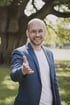 Lächelnder Mann mit Brille und blauem Sakko streckt im Park seine Hand zum Handschlag entgegen.